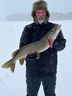 Man holding a large Northern Pike on a frozen lake with snow falling.